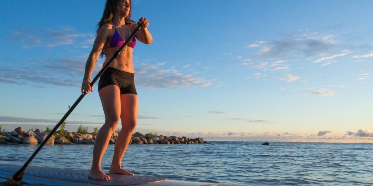 Female paddleboarding at sunrise with an oar in her hand, gliding across the calm water with the shore behind her