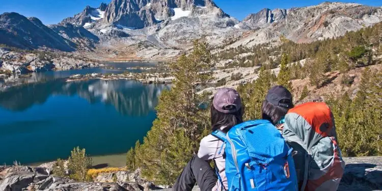 Two girls wearing backpacks sit on a mountain. They look toward the horizon at the eastern Sierra Nevada mountains and a lake.