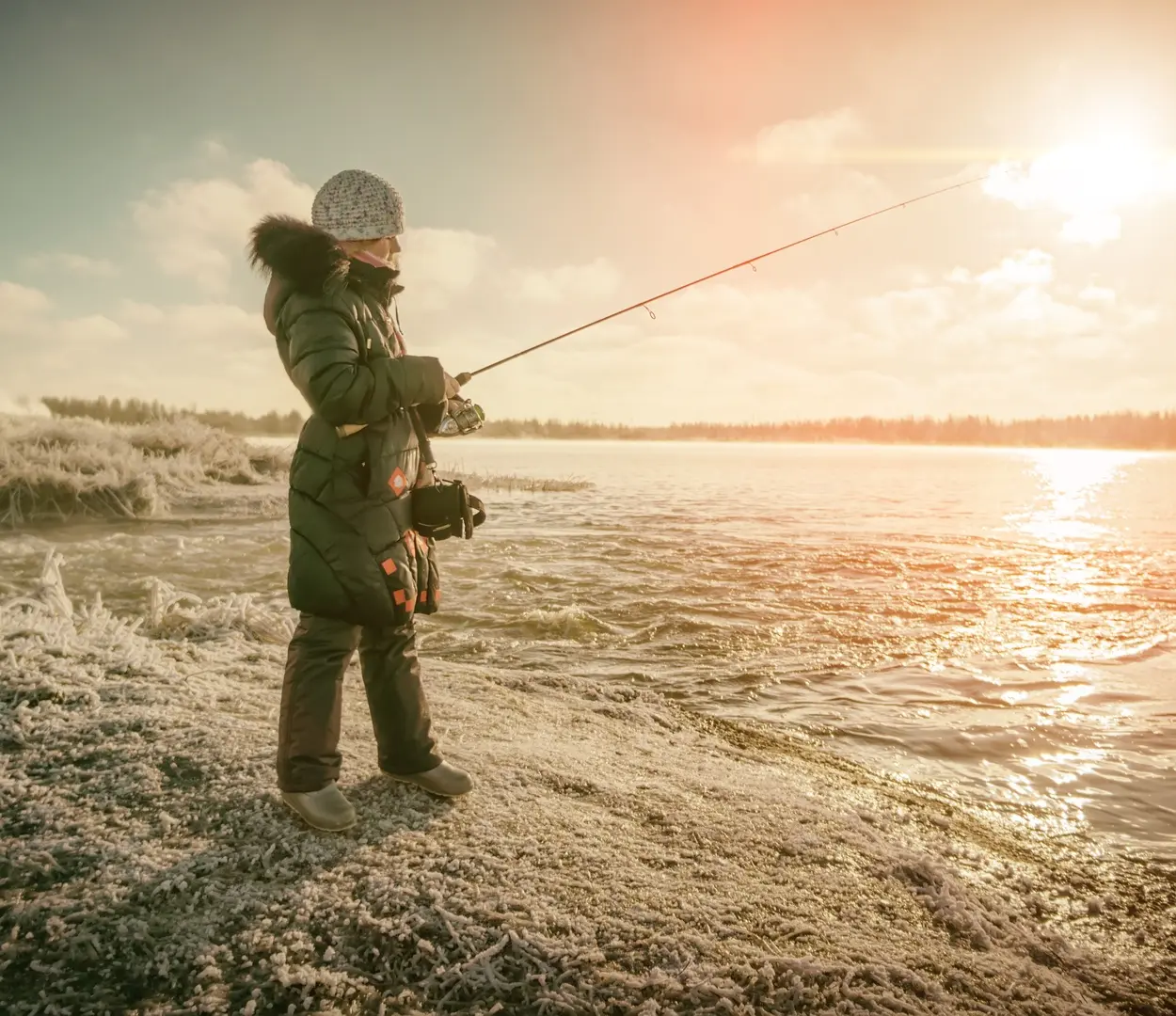 A woman wearing a long coat with a fur hood holds a fishing rod over a body of water. Snow sparsely covers the bank.
