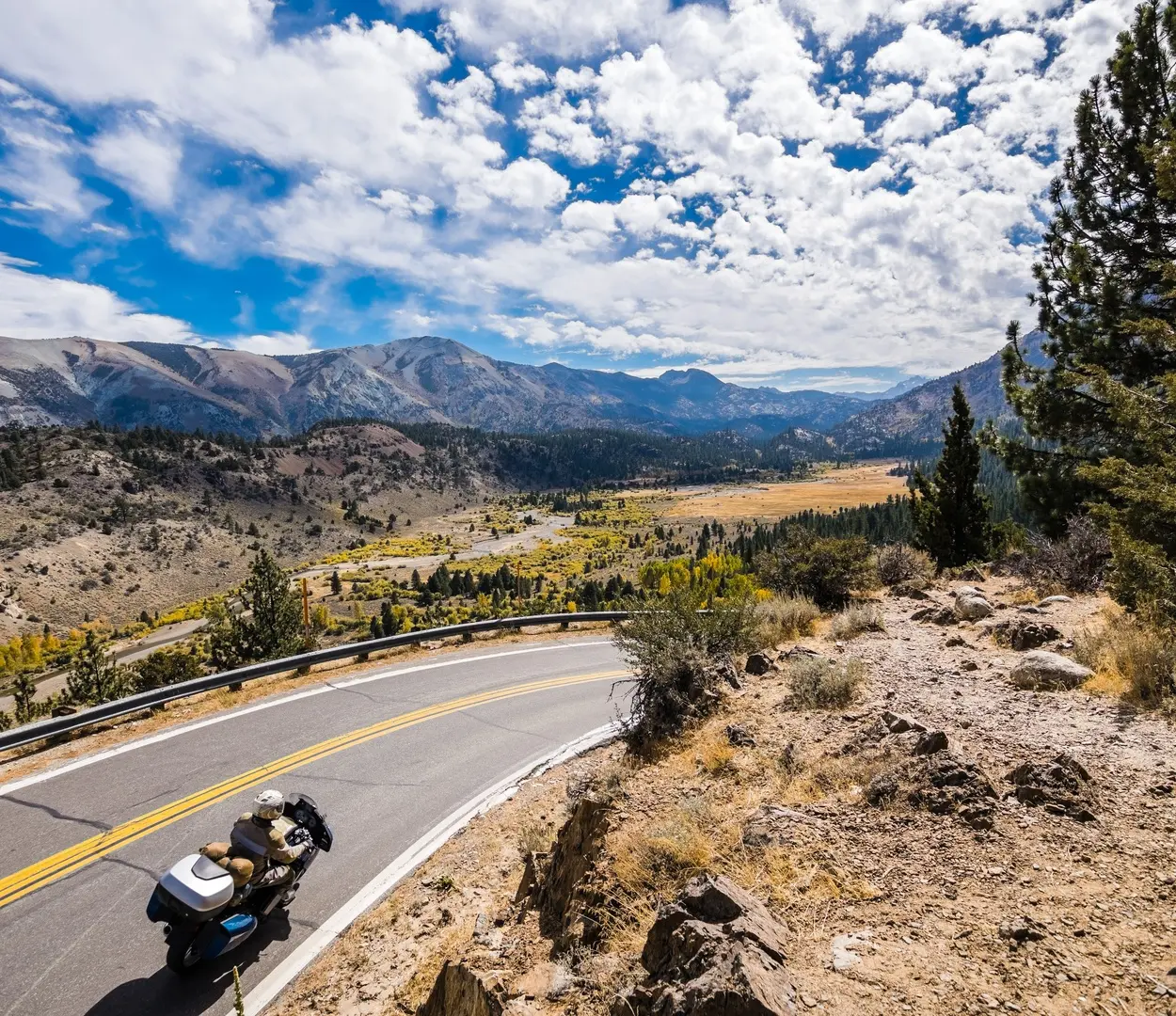 A motorcyclist travels down a well-kept, two-lane road in a high-altitude valley. Mountain ridges line the horizon.