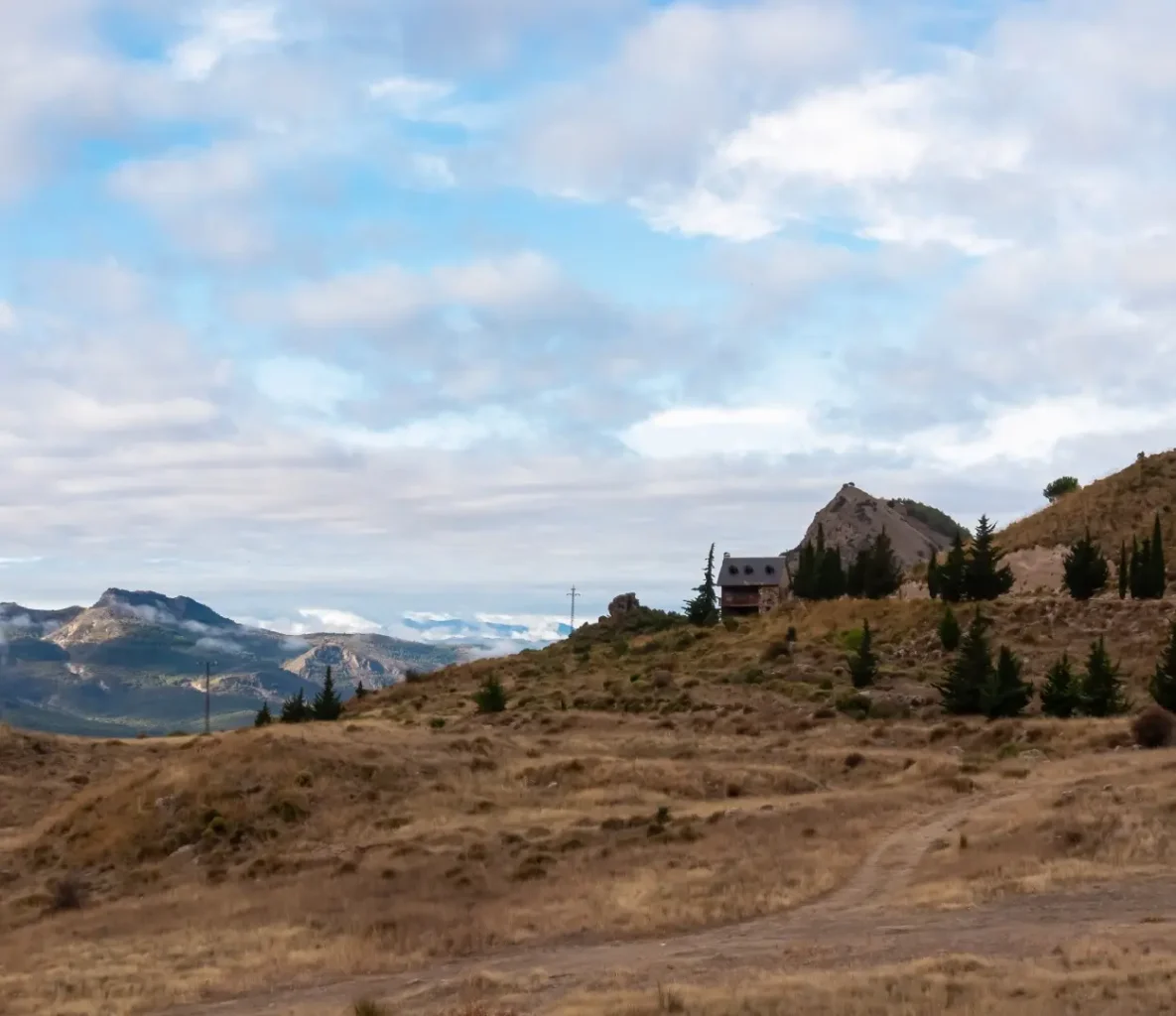 A small ultra-rural cabin sitting atop a hill covered in brown grass and small trees, overlooking a mountain range.