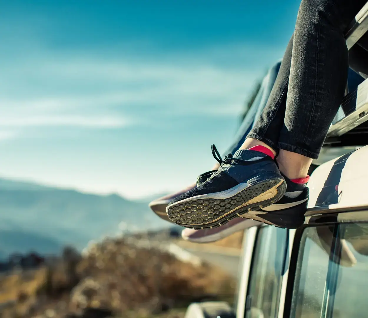 People sitting on the roof of their vehicle with their feet hanging off the side. There are some mountains in the background.