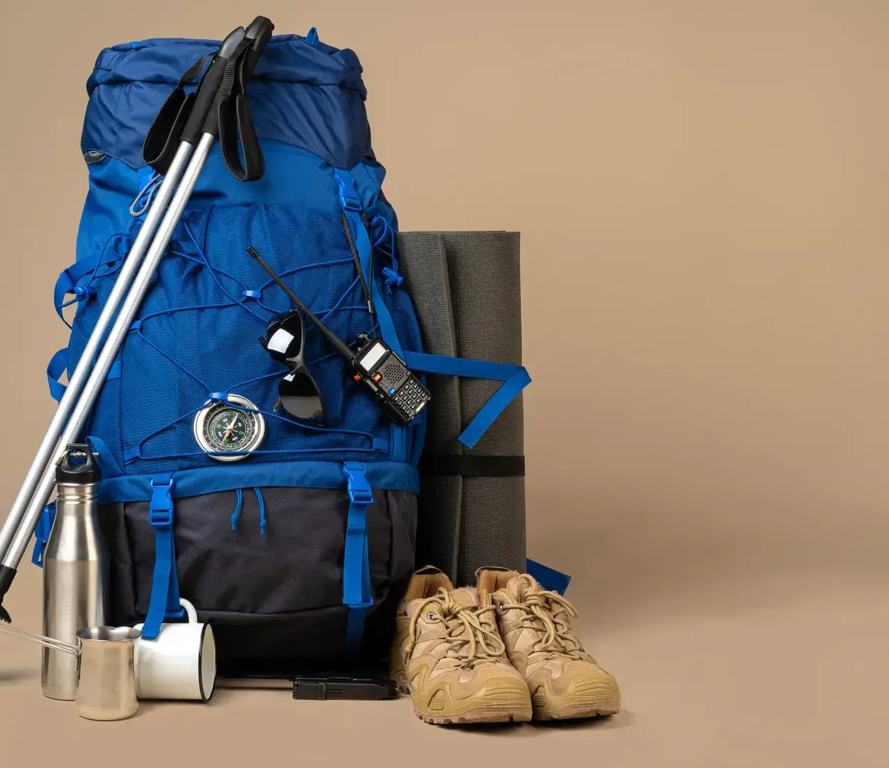 A large, blue backpack sits next to hiking books, hiking poles, a metal water bottle, and a white coffee mug.