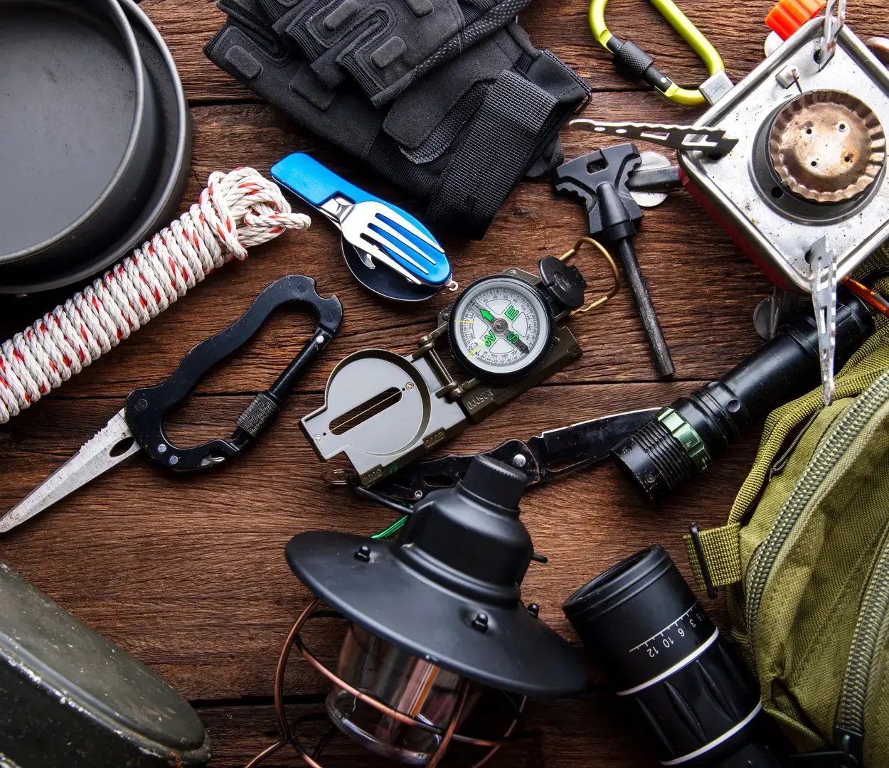 Trail gear and various tools spread across a wooden table next to a green backpack and a black cooking pot.