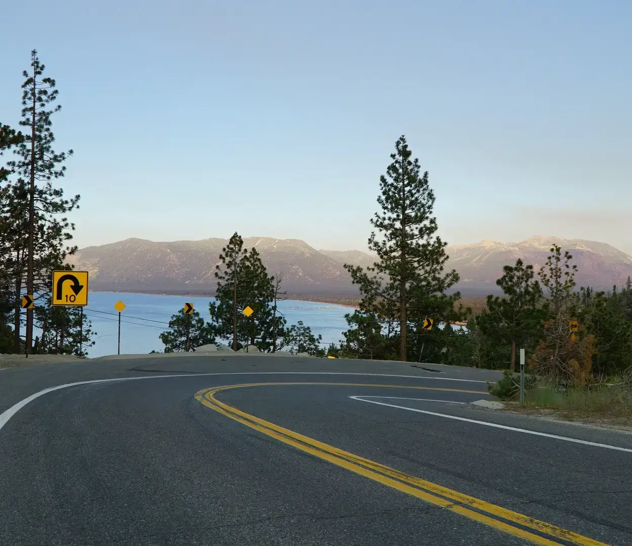 A curving mountain road descends toward a lake with pine trees, warning signs, and distant mountain peaks.