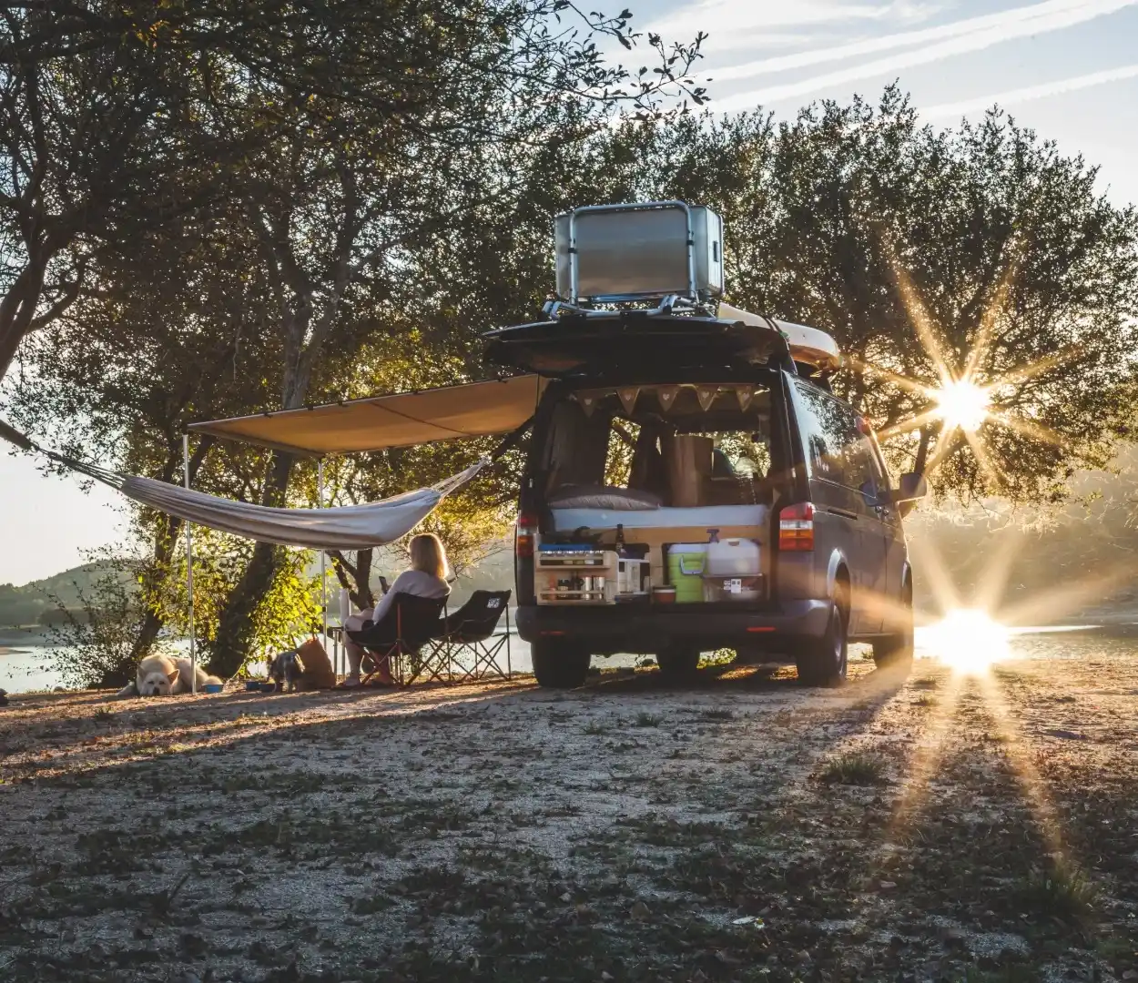 A camper van parked under trees. The van has an awning and a seating setup, while a woman sits and looks at the river.